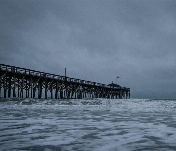 Ominous grey clouds hover over an old-looking oceanic pier. Dark, turbulent waves can be seen in the foreground.